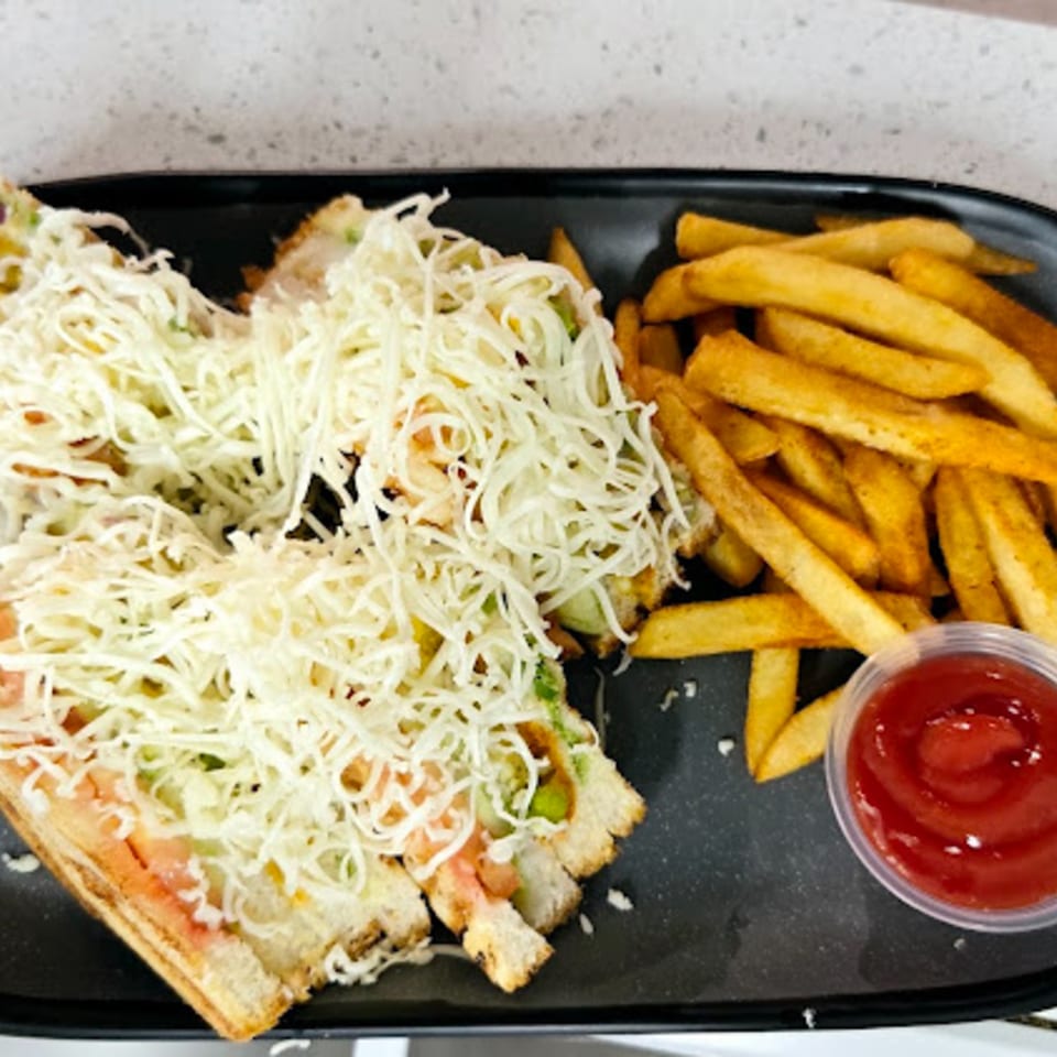 Indian vegetarian sandwich with shredded cheese, served with golden fries and ketchup on a black plate.