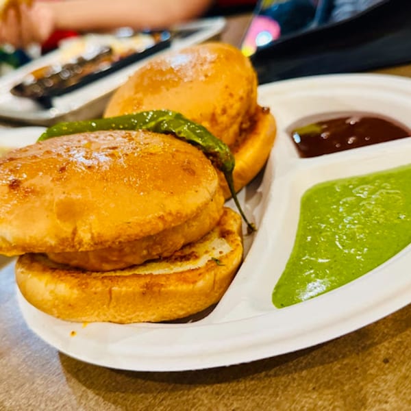 Two Indian vada pavs with a green chili, green chutney, and brown chutney on a white plate.