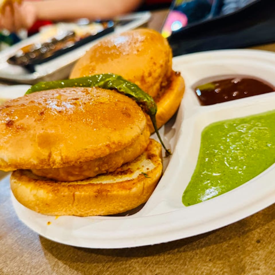 Two Indian vada pavs with a green chili, green chutney, and brown chutney on a white plate.